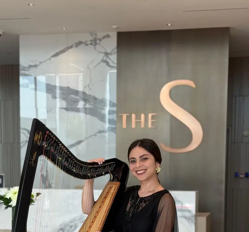 Harpist in a black gown standing beside a dark wood harp in a hotel lobby