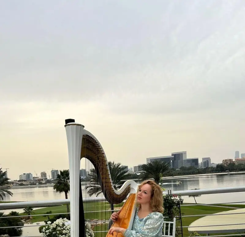 Harpist in a sequinned gown playing a white harp on an outdoor terrace by the water
