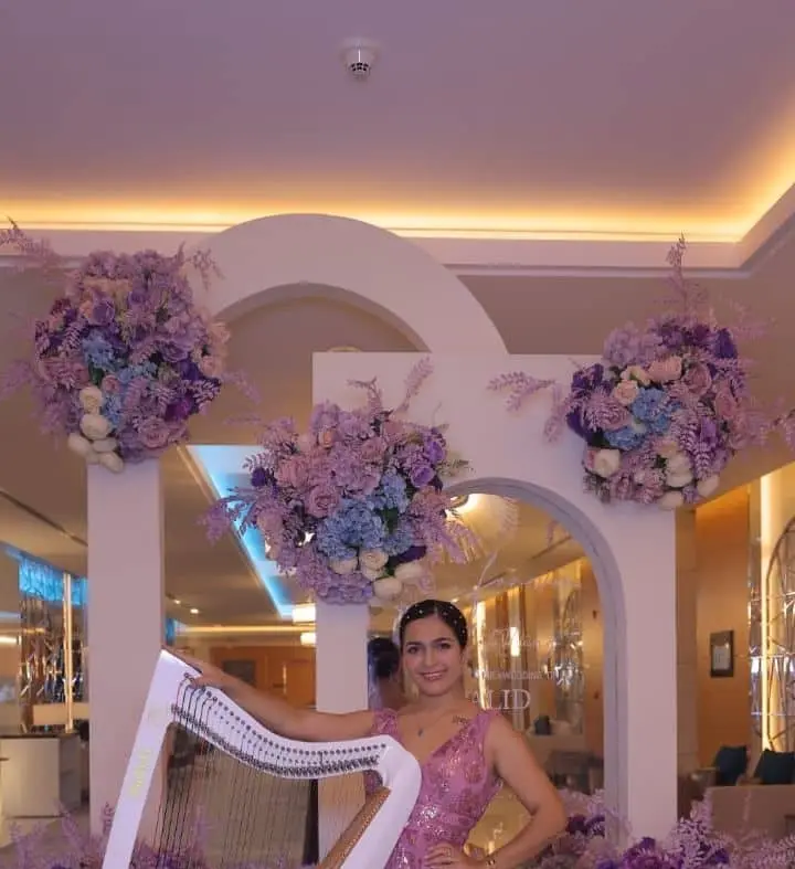 Harpist in a sequinned pink gown with a white harp under floral arches at an event