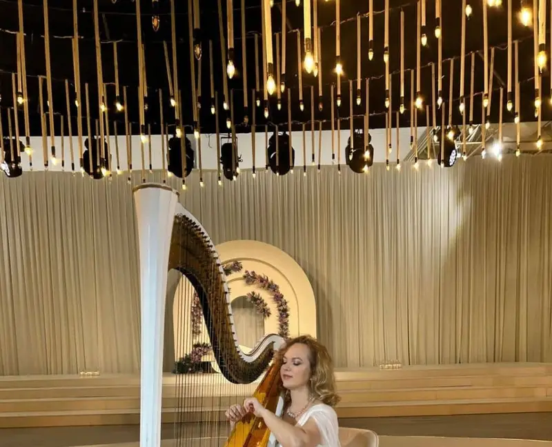 Harpist in white playing a white and gold concert harp in a ballroom with hanging lights
